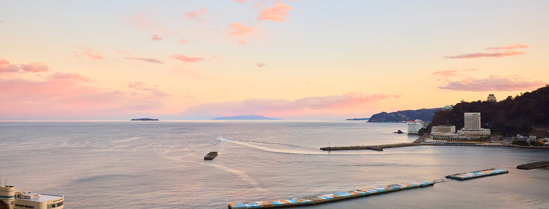 港のパノラマビュー。夕暮れ時で、空はピンクとオレンジに染まっている。港には複数の建物が立ち並び、沖合には小さな島影が見える。海面には船が航行した跡の波紋が広がっている。