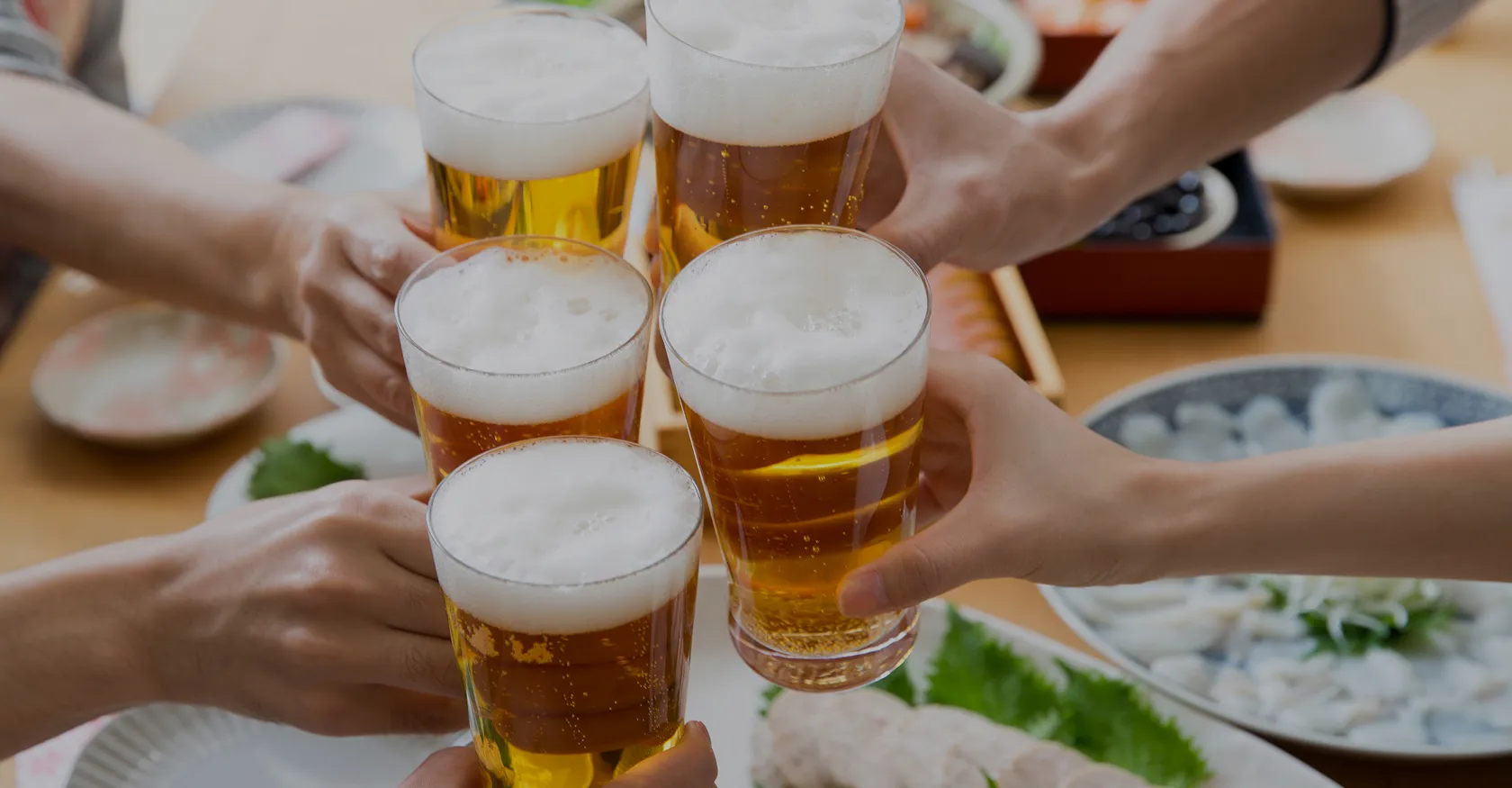 テーブルを囲んだ数人が、泡立つ黄金色のビールジョッキを掲げて乾杯しているクローズアップ。