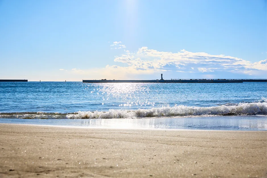 陽光きらめく穏やかな海辺の風景。手前には太陽の光を受けて輝く砂浜が広がり、波打ち際には白く砕ける波が優しく打ち寄せている。