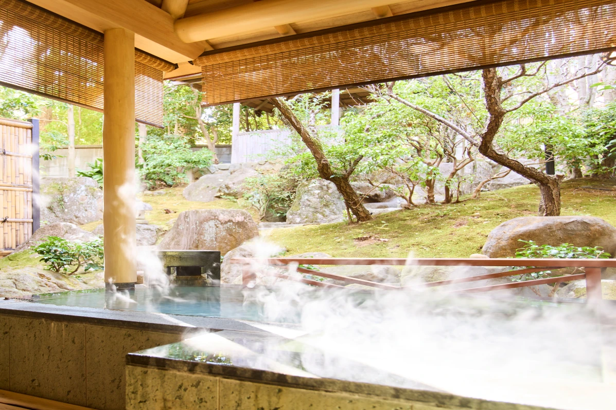 湯けむりが立ち上る、風情ある露天風呂の全景。浴槽の向こうには青々とした苔に覆われた日本庭園が広がり、様々な形の岩や木々が配されている。
