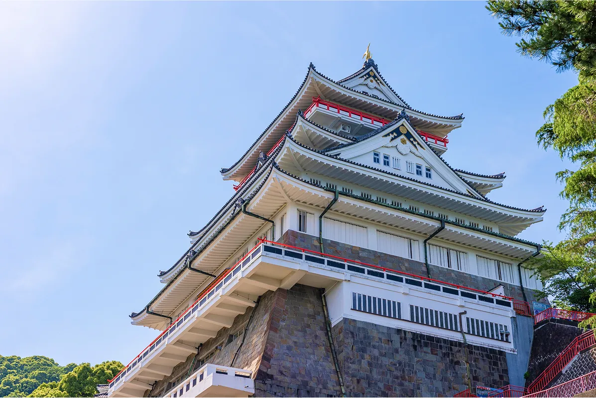 青く澄み渡る空を背景に、威厳ある姿でそびえ立つ城の天守閣をローアングルから捉えた一枚。白漆喰の壁と黒い瓦が織りなすコントラストが美しく、朱色の欄干がアクセントになっている。