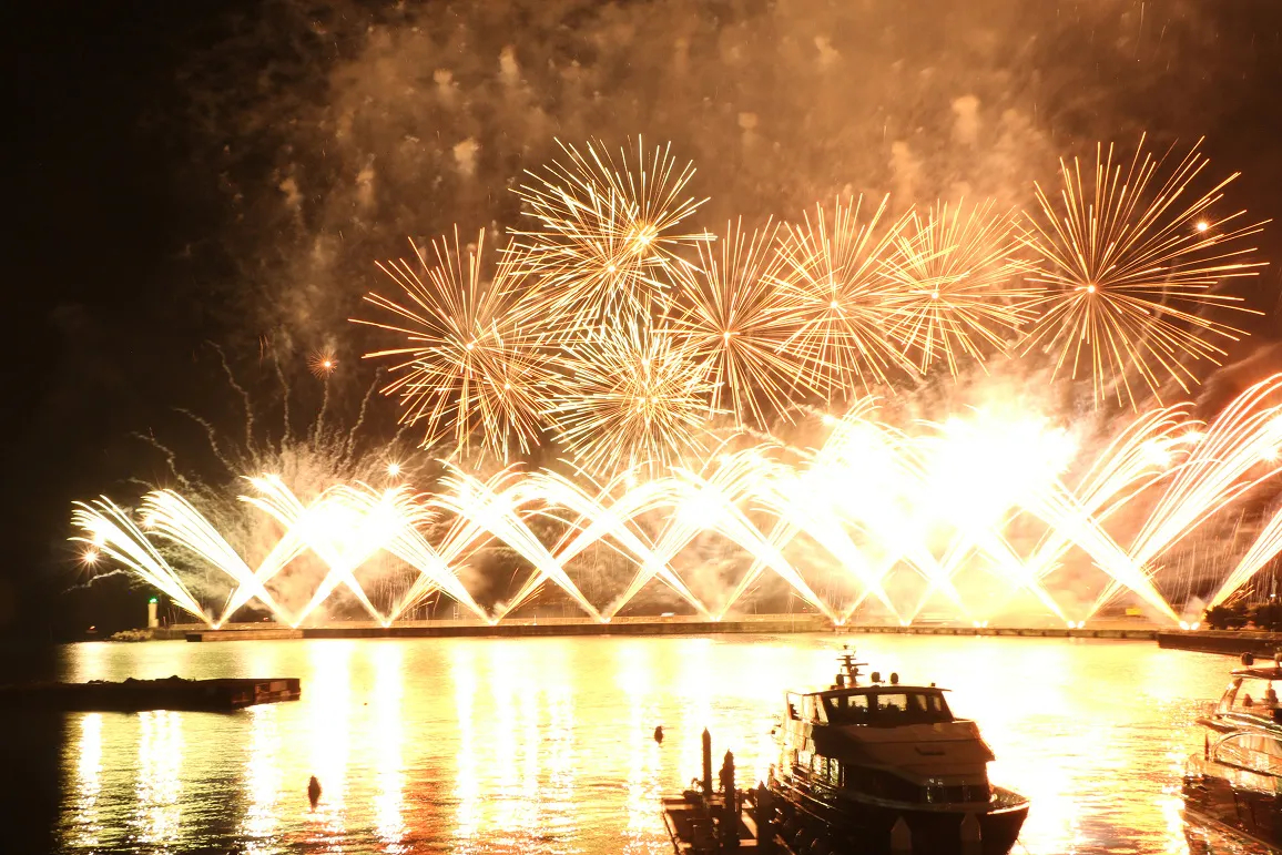 夜空に高く打ち上がる金色の花火と、水面を彩る横一列に並んだ扇状の水中花火。水面に光が反射し、手前には停泊中の船が見える。