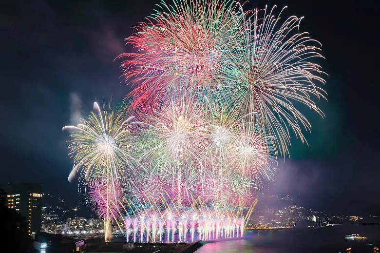 夜空に大きく打ち上がる、赤、緑、ピンク、白など色とりどりの花火。その下には、水面近くで一斉に放たれる連発花火が広がり、夜景を背景に幻想的な光景を作り出している。