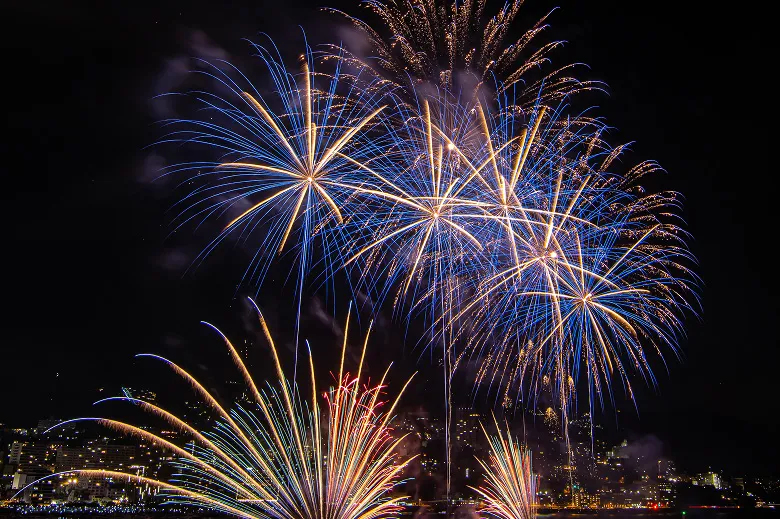 夜空に大きく打ち上がる、青色の光が放射状に広がる花火と、その下に広がる扇状の赤い花火。夜景を背景に、複数の花火が同時に夜空を彩る。