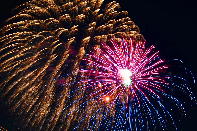 夜空に大きく打ち上がる、赤と青の鮮やかな光が広がる花火。その奥には、金色に輝く別の花火が重なり合って見える。
