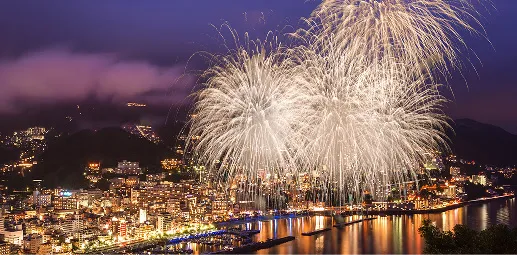 夜空を彩る盛大な花火が、街の灯りを背景に打ち上げられている。花火の光が水面に反射し、夜景と一体となっている。